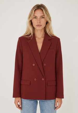 Donna bionda che indossa una giacca doppioppo rosso scuro e jeans azzurri, in piedi contro uno sfondo chiaro e semplice.