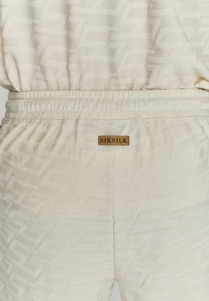 Jogging beige en tissu texturé avec un motif géométrique subtil. Taille élastique avec une étiquette de marque pour l'identification.