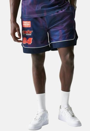 Person trägt lila und marineblaue Mesh-Sportshorts mit roten Logos und der Nummer 04, weiße Sportsocken und weiße Turnschuhe und steht vor einem schlichten Hintergrund.
