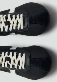 Par de zapatillas negras con cordones blancos y rayas blancas sobre fondo gris, vistas desde arriba.