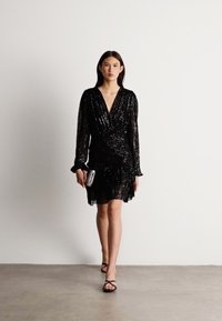 Vestido envolvente preto, com lantejoulas, mangas longas e bainha rodada. Acentuado com uma clutch brilhante e sandálias minimalistas com tiras.