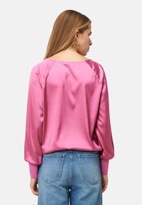 Blouse en satin rose à manches longues et col large, présentant une texture lisse et des coutures raglan aux épaules, associée à un jean bleu.