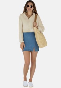 Hellbeige Langarmshirt, blauer Denim-Minirock mit vorderem Schlitz, gewebte sandfarbene Tragetasche, weiße Slipper, runde Sonnenbrille.