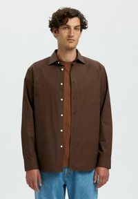 Braune Hemdbluse mit spitzem Kragen, ausgestattet mit weißen Knöpfen, kombiniert mit einem braunen Unterlayer, getragen mit blauen Jeanshosen.