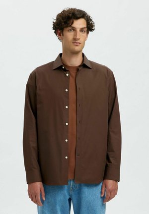 Camicia marrone con colletto a punta, caratterizzata da bottoni bianchi, abbinata a uno strato inferiore marrone e indossata con pantaloni in denim blu.
