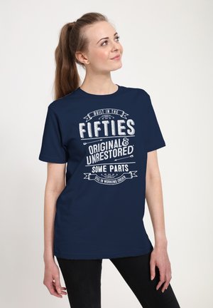 Kvinde med brunt hestehale iført navyblå t-shirt med teksten "Bygget i 1950'erne, original og urestaureret, nogle dele stadig i funktionsdygtig stand."