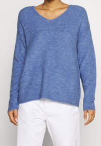 Personne portant un pull en maille bleu ample avec un col en V et des manches longues, associé à un pantalon blanc et des bagues dorées à la main droite.
