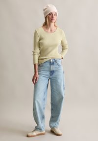 Haut à manches longues côtelé lime, jean large bleu clair, baskets beiges et bonnet crème, avec une silhouette décontractée et des textures douces.