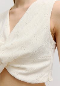 Weißes ärmelloses Top mit strukturiertem Spitzenmuster, V-Ausschnitt und gerafftem Detail an der Taille, mit einem kleinen Reißverschlussverschluss.