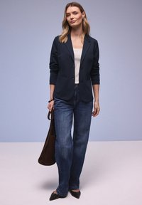 Femme portant un blazer noir, un haut blanc, un jean bleu, des chaussures noires, tenant un sac à main marron, debout devant un fond dégradé gris-bleu.