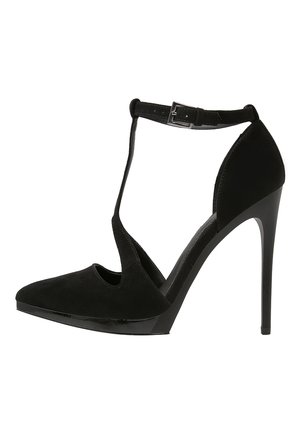 Chaussure à talon haut noire avec une finition en daim, dotée d'un bout pointu, d'un design à T-strap et d'une boucle de cheville ajustable. Semelle lisse et brillante.