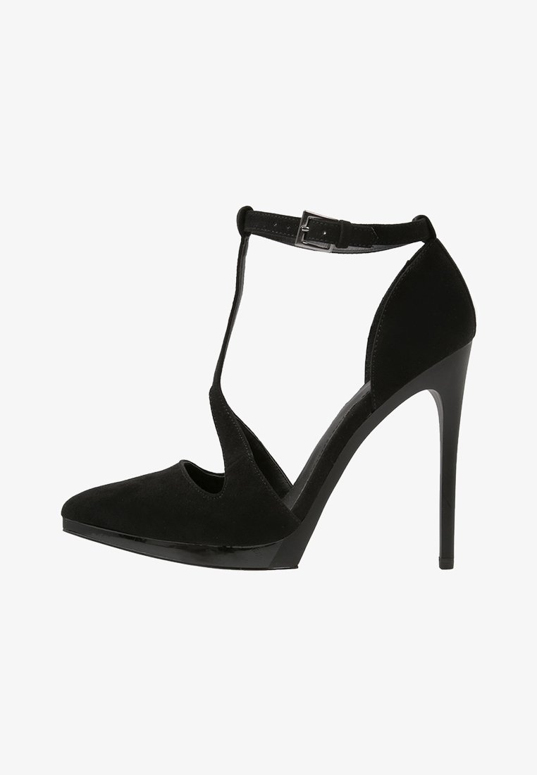 Chaussure à talon haut noire avec une finition en daim, dotée d'un bout pointu, d'un design à T-strap et d'une boucle de cheville ajustable. Semelle lisse et brillante.