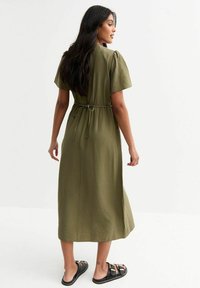 Vestido verde oliva de longitud midi con mangas cortas abullonadas y cintura ceñida con cinturón. Textura suave; se lleva con sandalias negras. Diseño sencillo, sin patrones.