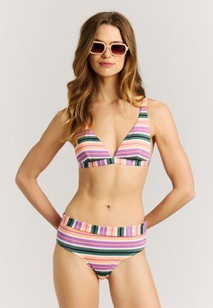 Žena v pásikových viacfarebných bikinách a svetlých veľkých slnečných okuliaroch, stojaca s rukami za chrbtom.