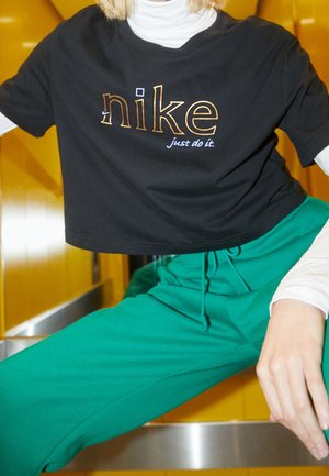 Osoba ubrana w czarny krótki t-shirt Nike na białej bluzce z długim rękawem i zielone spodnie, siedząca na tle żółtej ściany.