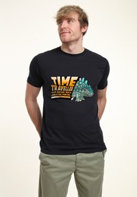Camiseta negra con un diseño gráfico de un dinosaurio estilizado con acentos turquesas y un texto en negrita que dice "VIAJERO EN EL TIEMPO DE OTRA DIMENSIÓN."