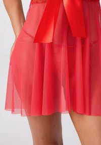 Czerwona przezroczysta siateczkowa narzutka z kokardą w talii. Charakteryzuje się rozszerzanym krojem oraz miękką, teksturowaną tkaniną. Długość sięga do połowy uda.