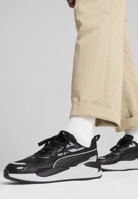 Svarta atletiska sneakers med texturerad mesh-ovandel, vit mellansula och svarta skosnören. Bärs med beige avsmalnande byxor och vita strumpor.