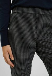 Pantalones de tejido con un patrón de pata de gallo en gris oscuro y azul marino, con una textura suave y una cinturilla ajustada. Mano descansando sobre los pantalones.