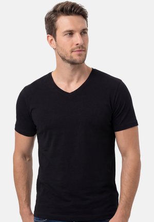 Junger Mann mit kurzen braunen Haaren, trägt ein schwarzes V-Ausschnitt-T-Shirt und blickt leicht nach rechts vor einem schlichten hellen Hintergrund.