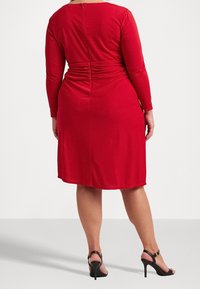 Červené šaty s dlouhým rukávem, s nařaseným detailem na zadní straně a kulatým výstřihem. V kombinaci s černými sandály na vysokém podpatku se řemínky.