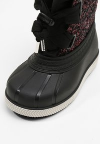 Close-up of black and white waterproof boot with textured sole, fabric upper with black and red pattern, and black laces tied in a bow.