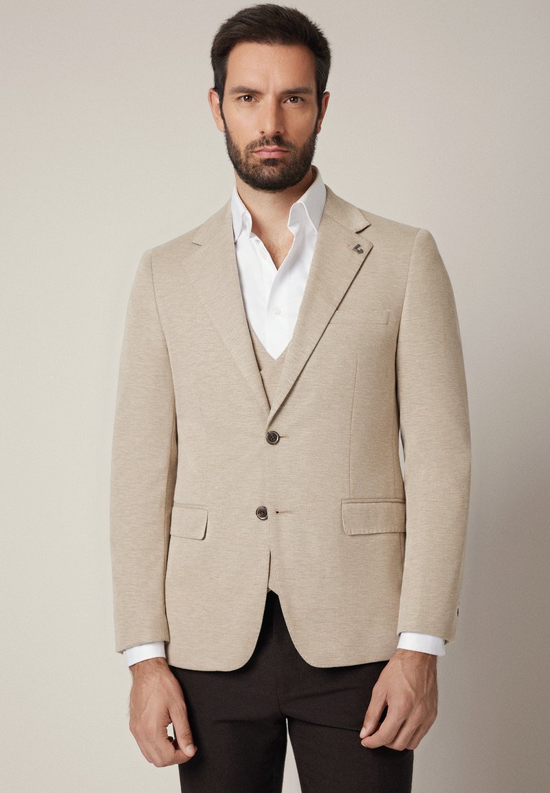 Mężczyzna w beżowej marynarce, dopasowanej kamizelce, białej koszuli i ciemnych spodniach stojący na neutralnym tle.