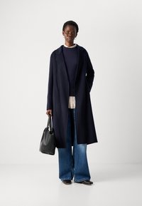 Mulher em pé com um longo casaco azul-marinho, suéter escuro, camisa bege, jeans de perna larga, sapatos pretos, e segurando uma bolsa de couro preta contra um fundo simples.