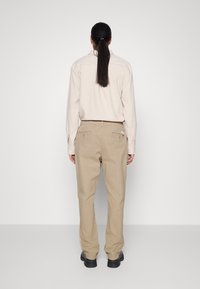 Beige katoenen broeken met een elastische tailleband en achterzakken, gecombineerd met een lichtbeige lange mouw shirt, gezien van achteren. Zwarte schoenen.