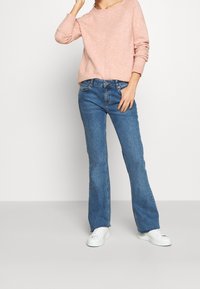 Maglione rosa chiaro in misto cotone con vestibilità comoda e scollo ampio, abbinato a jeans a zampa d'elefante blu e sneakers bianche.
