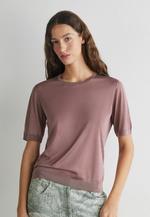 Frau mit langen braunen Haaren, die ein fliederfarbenes kurzärmeliges Strickoberteil und graue gemusterte Hose trägt, steht vor einer schlichten weißen Wand.