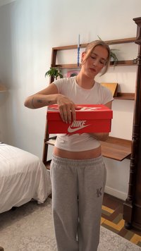 Rød Nike skoæske holdt med begge hænder, med det hvide Nike-logo. Værelsesindstillingen inkluderer en seng og træhylder i baggrunden.