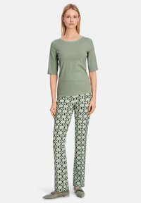 Grünes Kurzarmoberteil, gemusterte Hose mit geometrischem Design in Grün, Schwarz und Weiß, dazu passende flache Schuhe.