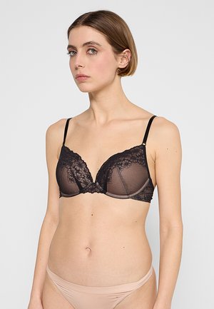 Młoda kobieta ubrana w czarny koronkowy biustonosz z fiszbinami i beżowe majtki z niskim stanem, stojąca na jednolitym jasnoszarym tle.