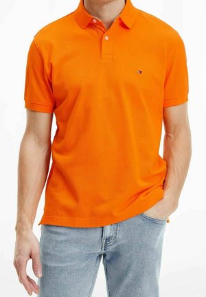 Man draagt een feloranje polo met korte mouwen en een klein logo, gecombineerd met lichtblauwe spijkerbroek, één hand in de zak, tegen een witte achtergrond.