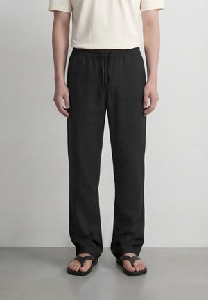 Personne portant un pantalon ample noir à cordon de serrage, une chemise blanche à manches courtes et des sandales noires ouvertes, debout sur un sol gris.