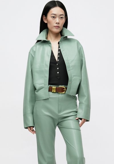 Jaqueta cropped de couro verde menta com um colarinho grande, combinada com uma blusa preta de botões e calças de couro verde menta combinando, acentuada por um cinto castanho.