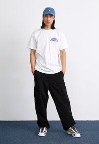 Camiseta branca com gráfico roxo, calças cargo pretas e boné azul. O conjunto apresenta um corte descontraído e ténis robustos com detalhes amarelos.