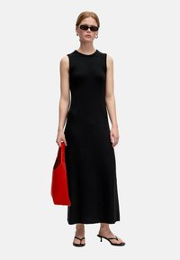 Schwarzes ärmelloses Kleid aus strukturiertem Strick, knöchellang. Die Model hält eine rote Tote-Tasche. Schwarze schmale Sandalen runden das Outfit ab.