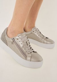 Sneakers argento metallici con pannelli laterali testurizzati, linguetta imbottita e chiusura con lacci. Presentano un dettaglio con zip sul lato e una suola bianca.