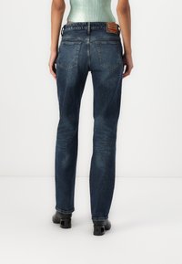 Mujer vestida con jeans de mezclilla azul oscuro y zapatos de tacón negros, de pie con la espalda hacia la cámara frente a un fondo blanco liso.