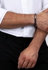 Marinblå flätad läderarmband med en metallisk detalj, bärs på ett handled. I bakgrunden syns en vit skjorta och svarta byxor.