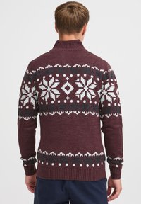 Maglione bordeaux con colletto alto, caratterizzato da motivi di fiocchi di neve bianchi e verde scuro. Polsini e bordo a costine, tessuto a maglia testurizzato.