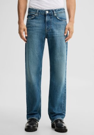 Mann trägt gerade geschnittene blaue Jeans, schwarze Schuhe und ein weißes T-Shirt und steht vor einem einfarbigen Hintergrund.