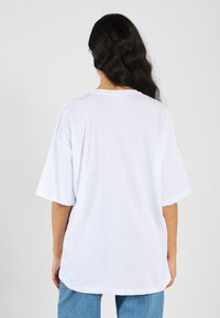 Bílý oversized T-shirt vyrobený z jemné bavlny, s kulatým výstřihem a krátkými rukávy, zobrazený ze zadní strany. Jednoduchý design, bez vzorů.