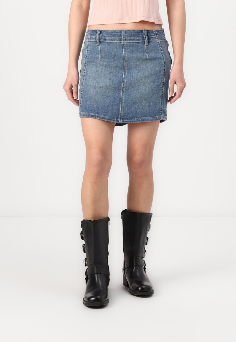 Jupe en denim de style mini avec une silhouette ajustée, de couleur bleu clair, poches avant et coutures. Associée à des bottes noires en cuir montantes avec des boucles.