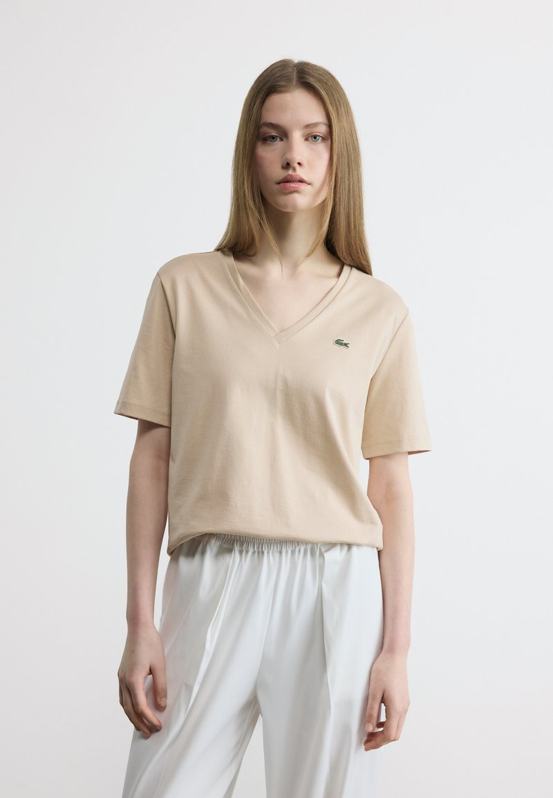 Νεαρή γυναίκα με ίσια μαλλιά φοράει μια μπεζ μπλούζα με V λαιμόκοψη και λευκό παντελόνι με ελαστική μέση, στέκεται μπροστά σε ένα απλό λευκό φόντο.