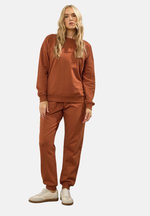 Vrouw staand, gekleed in een roestkleurige sweatshirt en bijpassende joggerbroek met witte sneakers tegen een effen achtergrond.