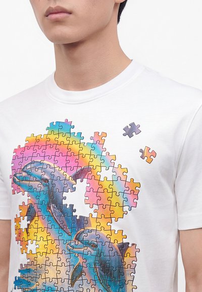 Άτομο που φοράει λευκό T-shirt με πολύχρωμο σχέδιο παζλ δύο δελφινιών και μερικά κομμάτια παζλ που λείπουν.