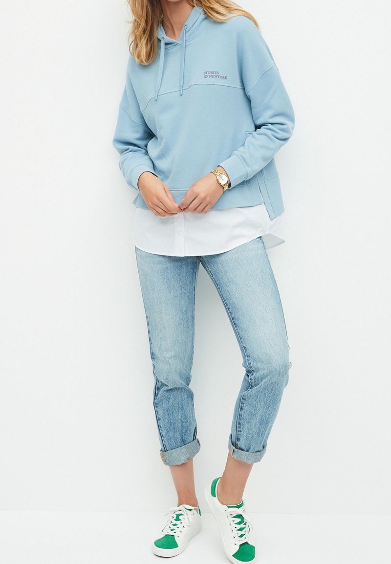 Sudadera de color azul claro con bolsillo frontal y el texto "HISTORIAS DE NUEVA YORK", superpuesta sobre una camisa blanca de botones, combinada con jeans claros y zapatillas deportivas.